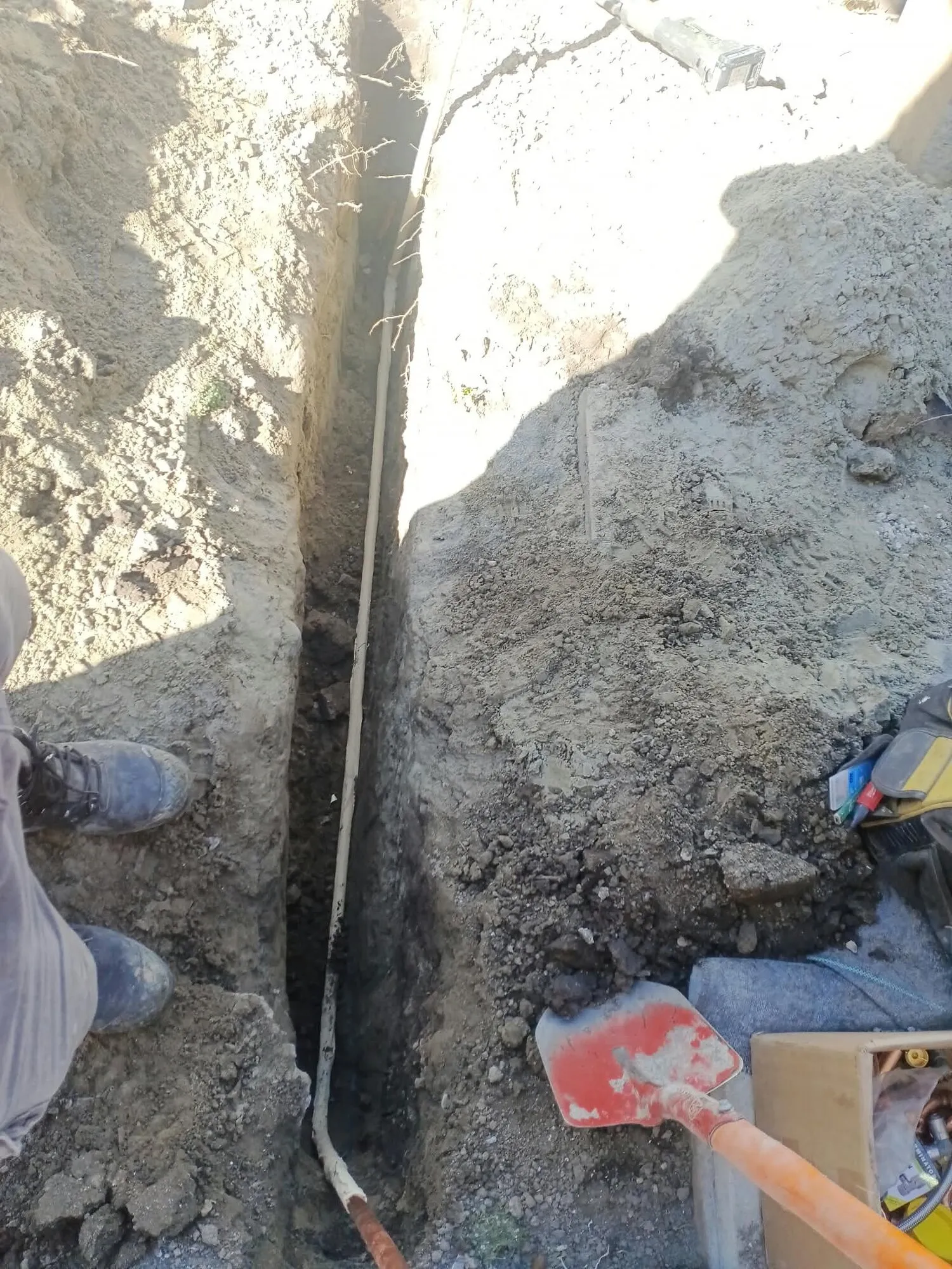 A new drainage line sitting in the bottom of a narrow hand dug trench, with a drainlayer standing in the trench behind it
