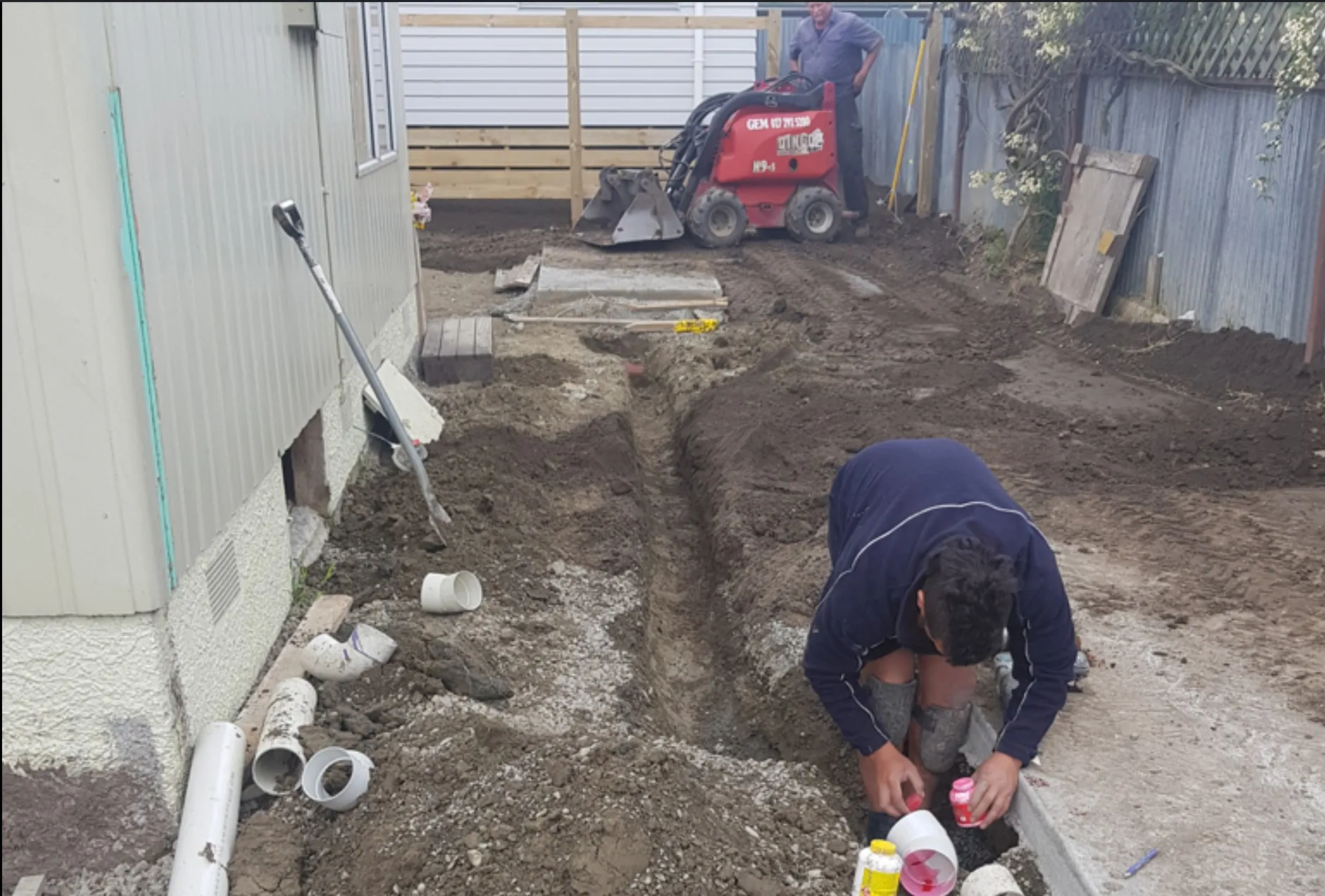 New drainage part laid in an open trench beside a Napier house, with PVC pipe and fittings on the ground and a mini loader behind