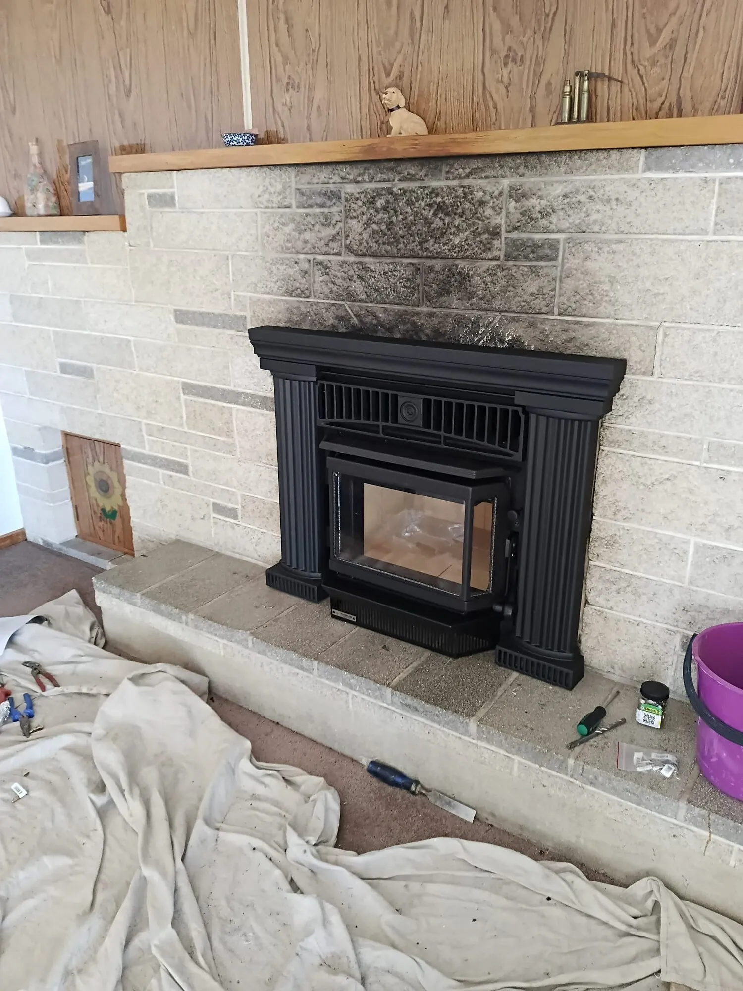 A black inbuilt wood fire with fluted columns fitted into a stone fireplace on a raised hearth, drop cloths and tools still on the floor at the end of the job