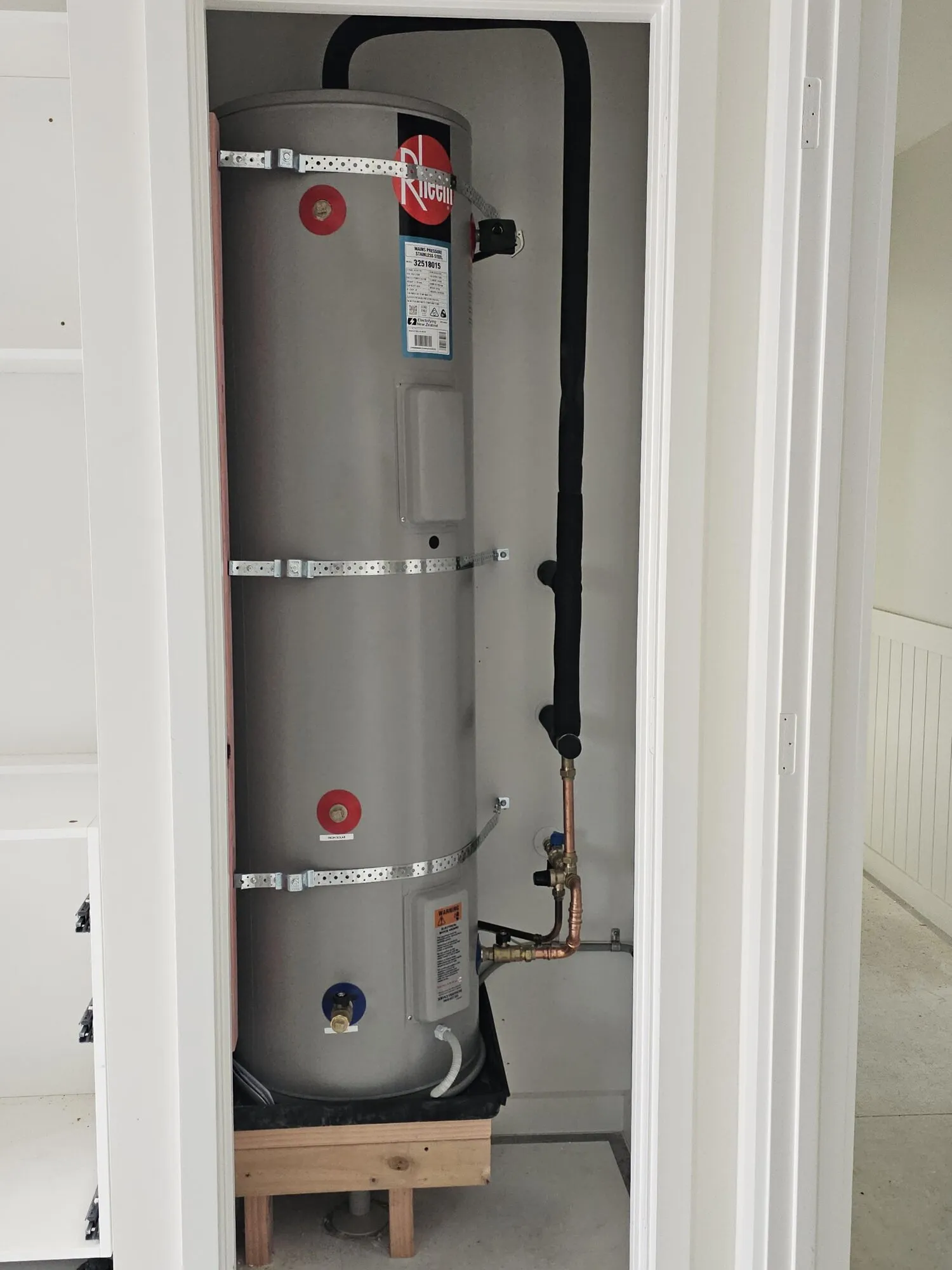 A Rheem hot water cylinder installed in a hallway cupboard, strapped and sitting on a timber plinth with the pipework run tidily behind it