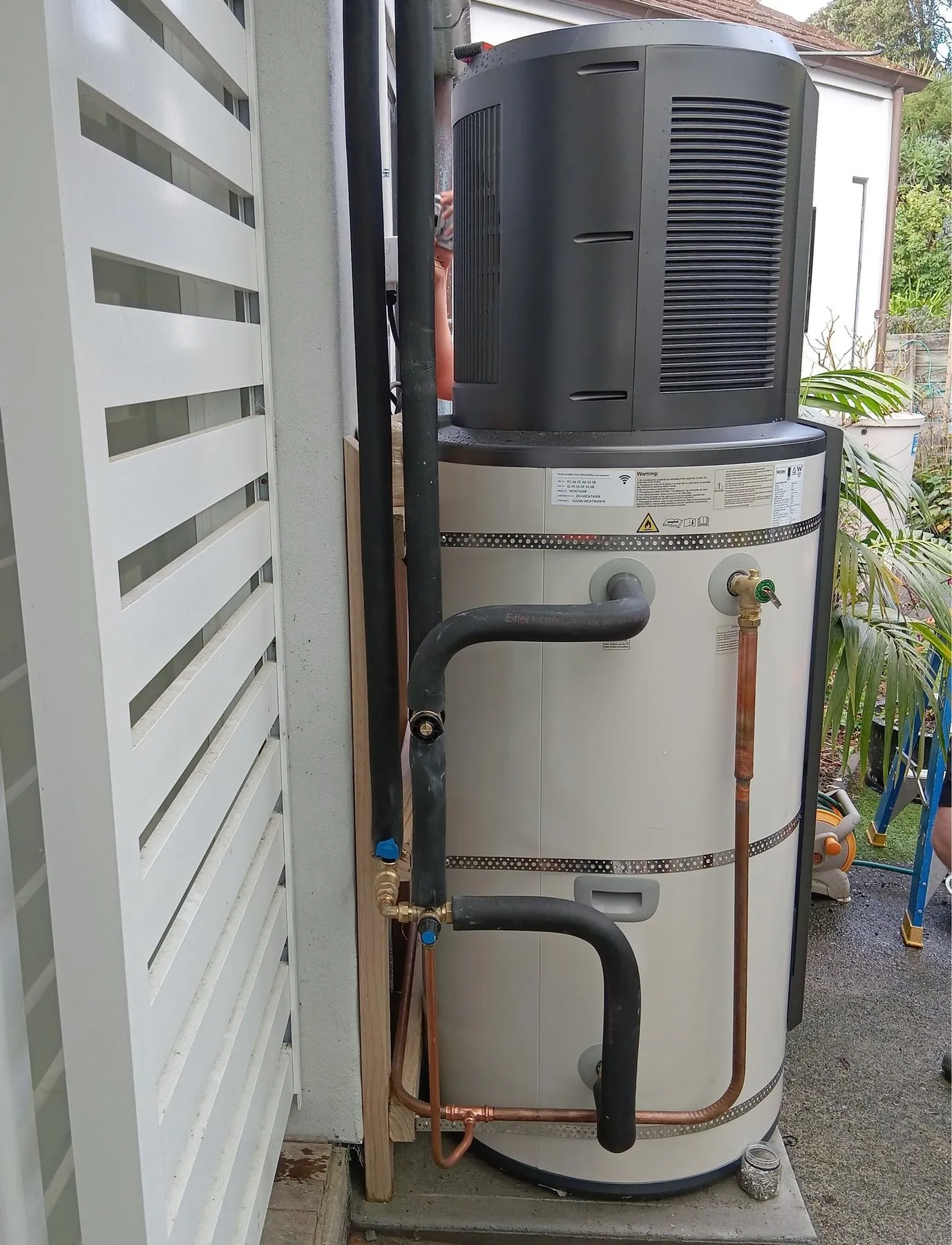 A heat pump hot water cylinder installed on the side of a Napier house, with lagged pipework and a tempering valve