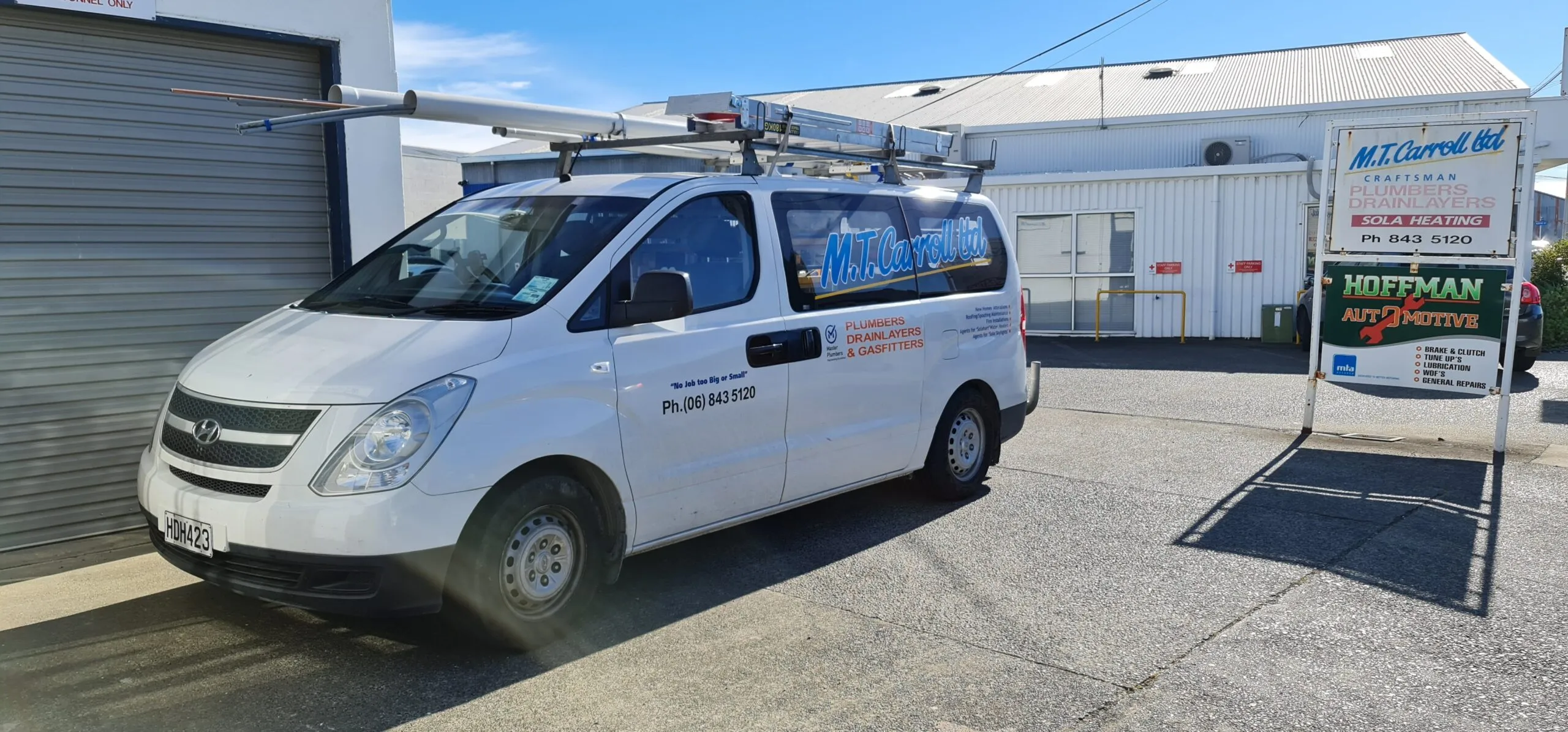 An M.T. Carroll Limited van loaded with ladders and lengths of pipe on the roof rack, outside the workshop in Onekawa