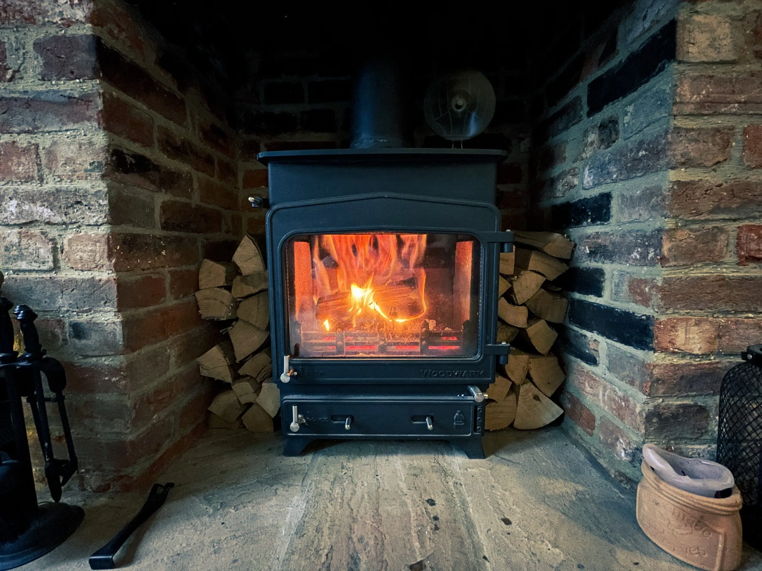 A wood burner alight in a brick fireplace opening, with split firewood stacked either side
