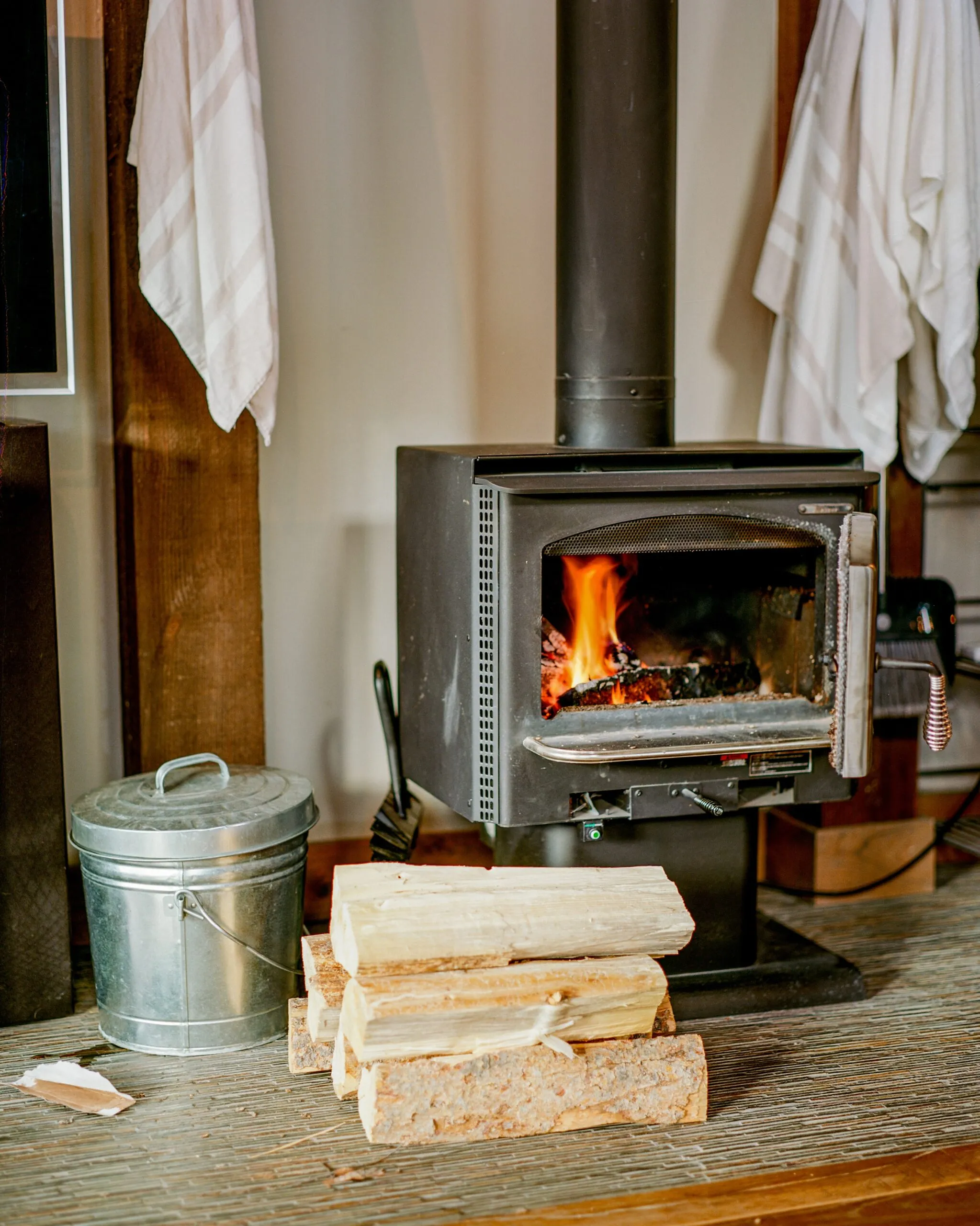 A freestanding wood burner alight on its hearth, flue running up to the ceiling, with split logs and an ash bucket beside it